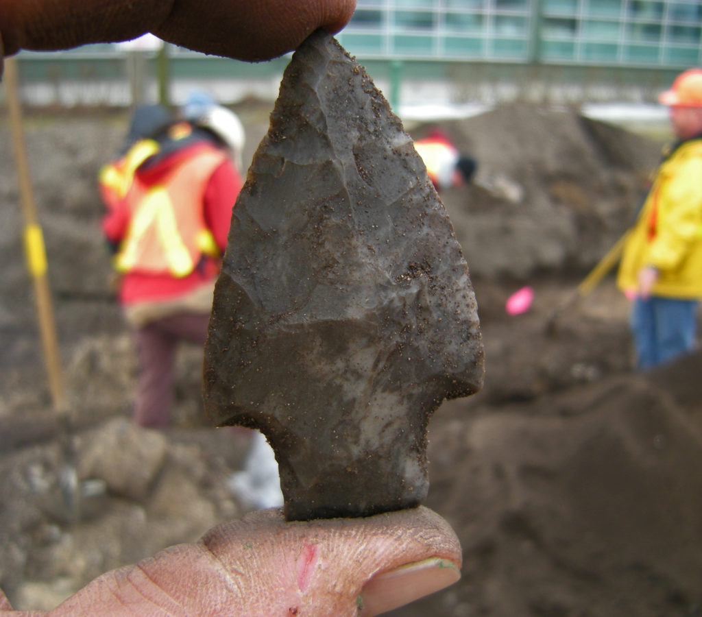 A stone arrowhead from the Peace Bridge site held between two fingers, with an excavation in the background
